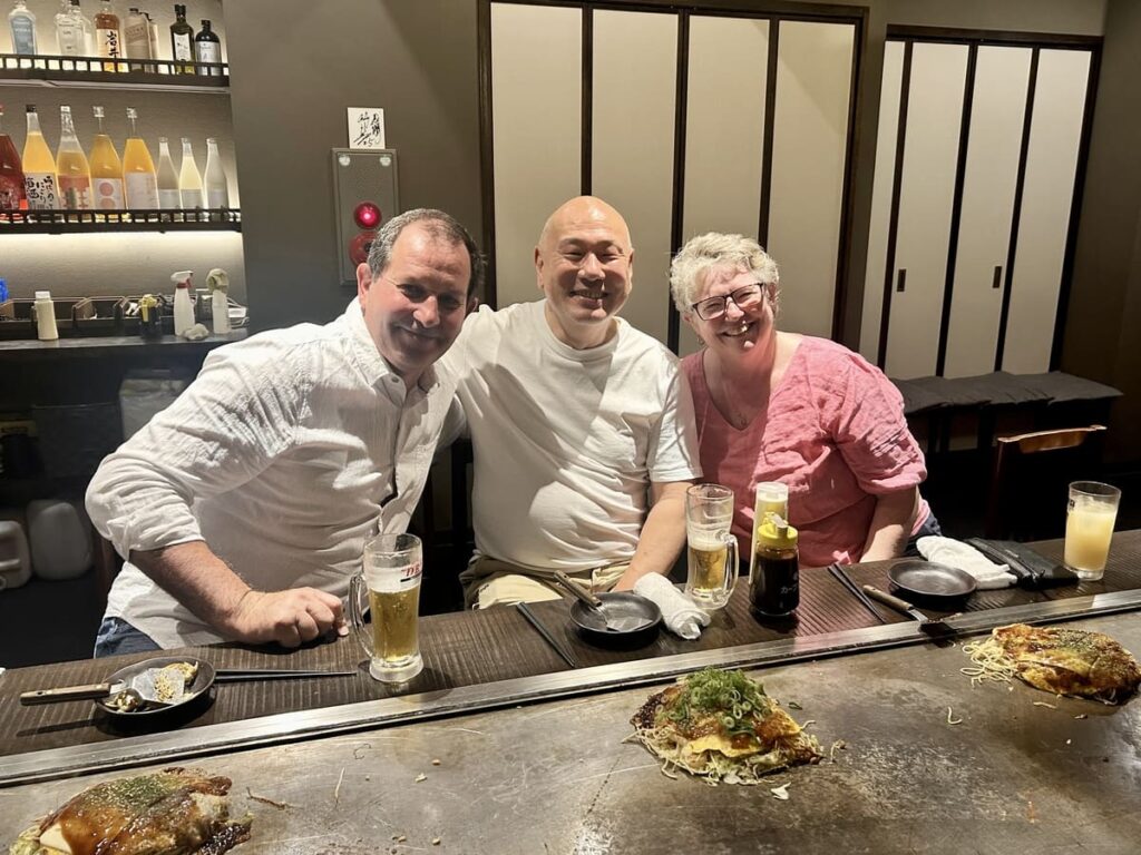 Three smiling people posing together behind a teppan griddle counter inside an Okonomiyaki restaurant, with freshly cooked Japanese savory pancakes and glasses of beer in front of them.
