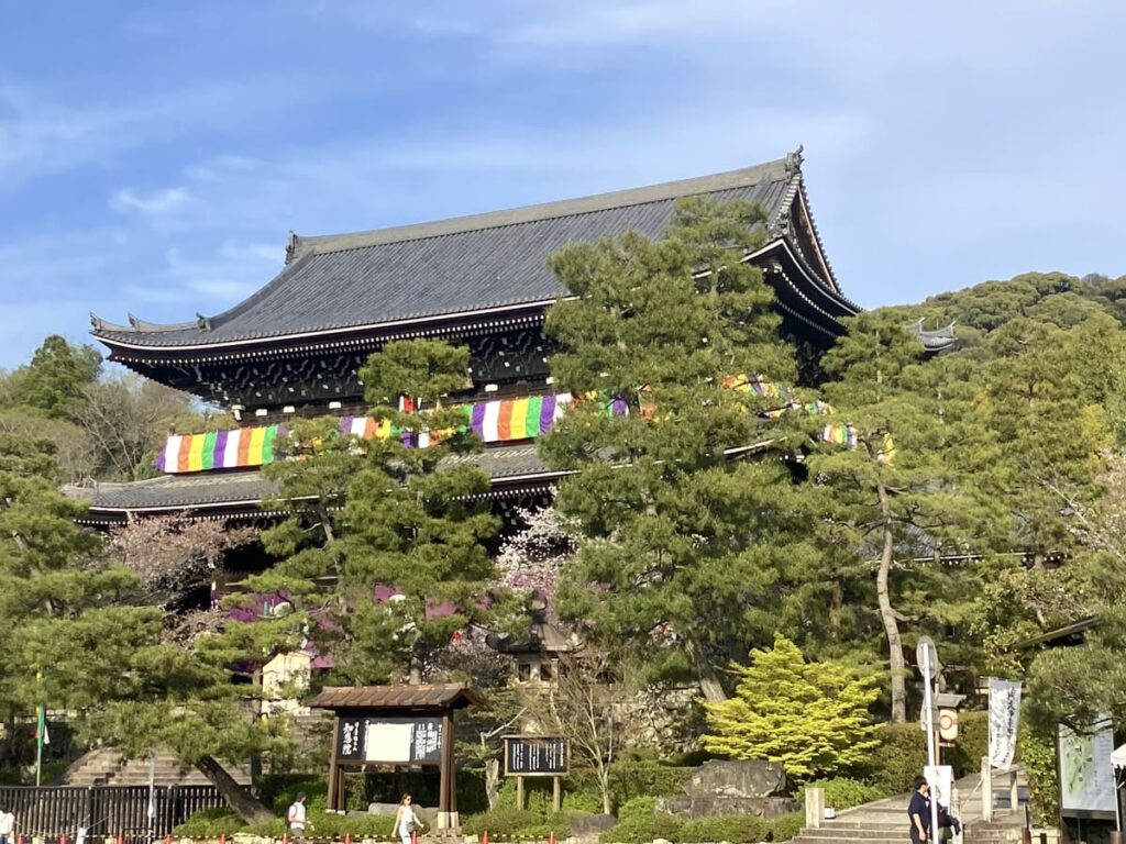 The massive, historic wooden Sanmon Gate at Chion-in Temple in Kyoto, adorned with colorful five-colored Buddhist banners and surrounded by pine trees.