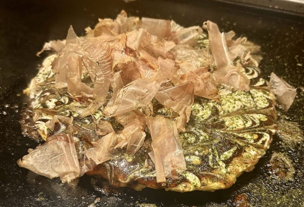 Okonomiyaki, a Japanese savory pancake topped with dancing bonito flakes cooking on a teppan grill.