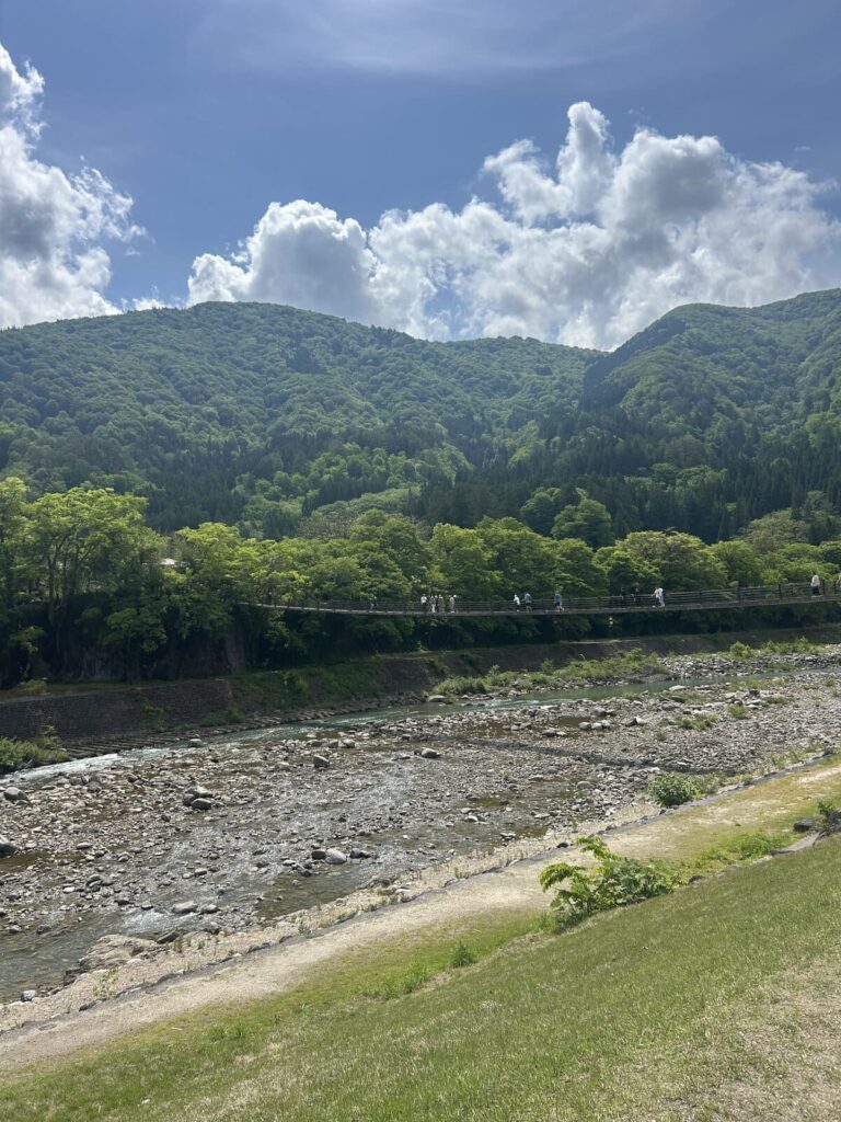 A long pedestrian suspension bridge crossing a rocky riverbed, surrounded by lush green forested mountains under a bright cloudy sky with visitors walking across.